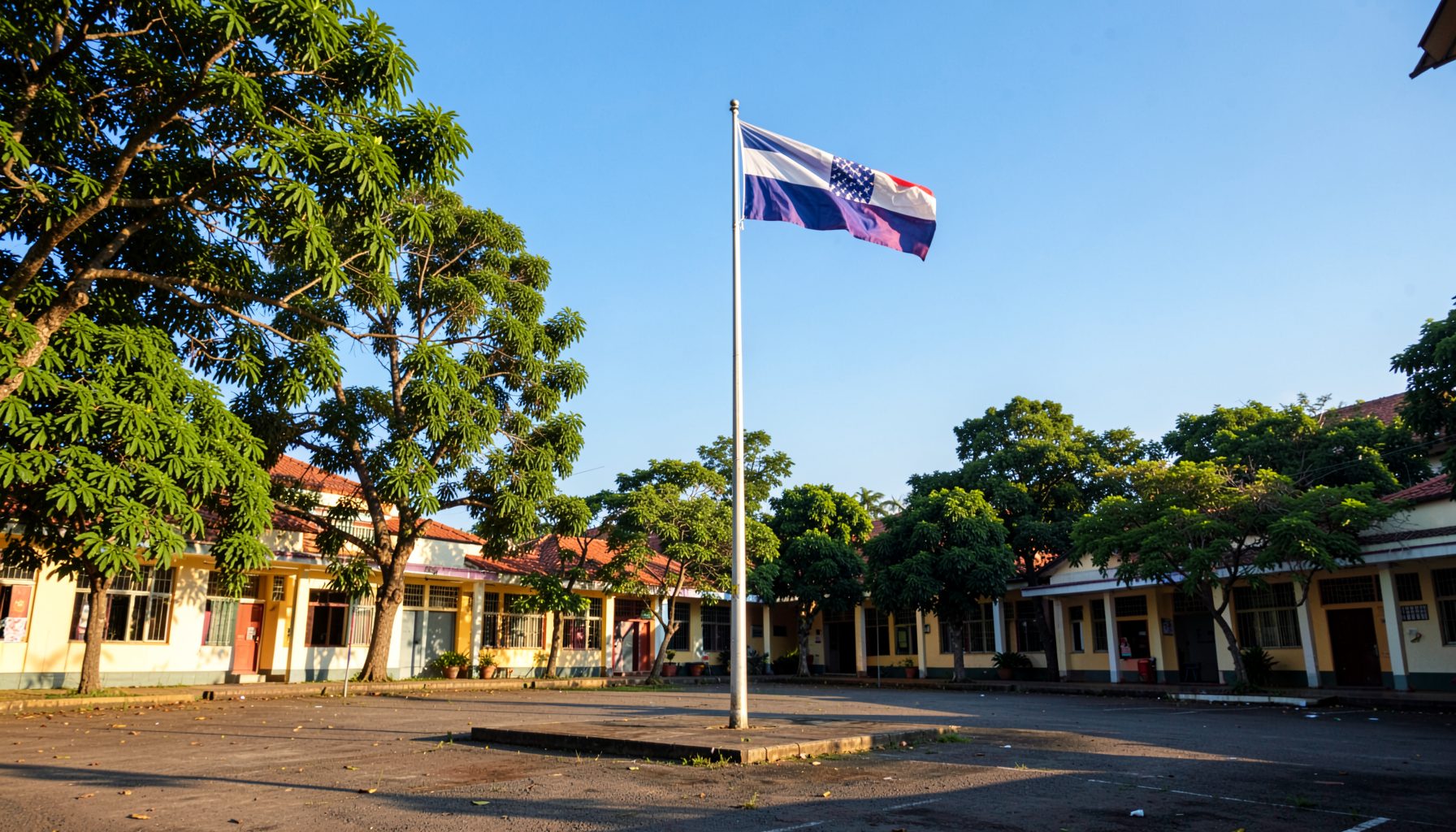 Wide view of a school campus courtyard with flagpole and Philippine flag against a bright blue sky, surrounded by greenery